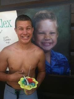 Smiling shirtless man holding a colorful shaved ice in front of a large child portrait