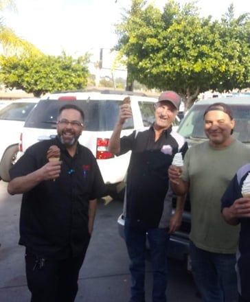 Three smiling people standing outdoors by a white SUV, one holding a microphone and another raising a fist.