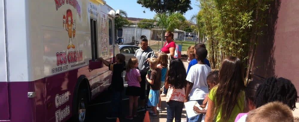 Children line up beside a pink food truck on a sunny street.