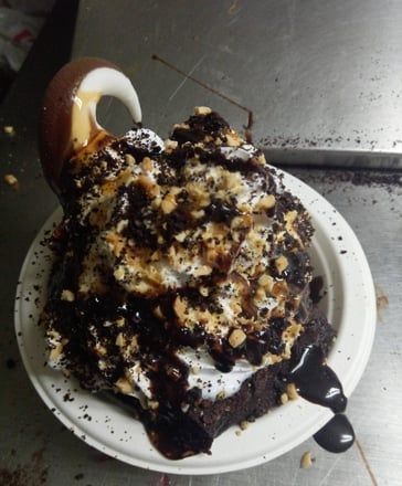 Dessert topped with whipped cream and chocolate syrup on a white plate, with a spoon beside it