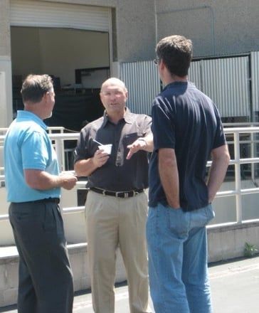Three men standing and talking outdoors by a building entrance.