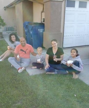Five people sitting on a lawn outside a garage, holding drinks and smiling.