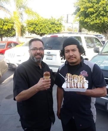 Two men standing outdoors holding drinks and a tray of cupcakes, with parked cars and trees behind them.