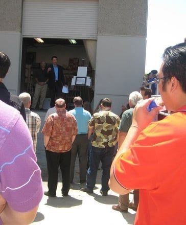 People gathered outside a warehouse-style building, facing an open doorway with a man speaking inside.