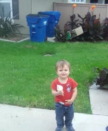 Toddler in a red shirt holding an ice cream cone on a driveway with a grassy yard and blue recycling bins.