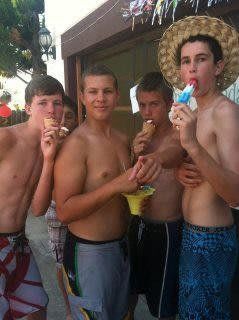 Four shirtless friends outdoors, smiling and eating ice pops by a building.