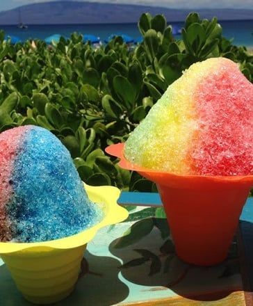 Two colorful shaved ice desserts in yellow and red cups by the ocean and greenery