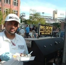 Vendor holding a plate of food beside a barbecue grill and BIG DEV’S sign at an outdoor market