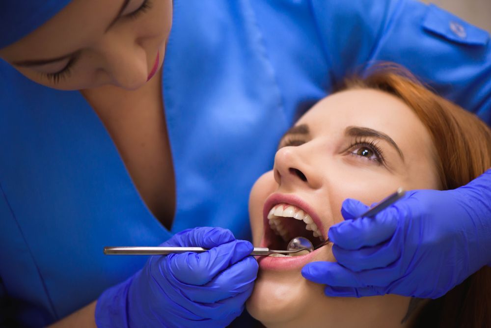 Dentist examining a patient's teeth with tools, both wearing blue gloves and scrubs, white interior.