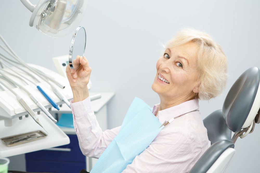 Woman at dentist's office smiling, looking at her teeth in a mirror. Dental tools are in background.