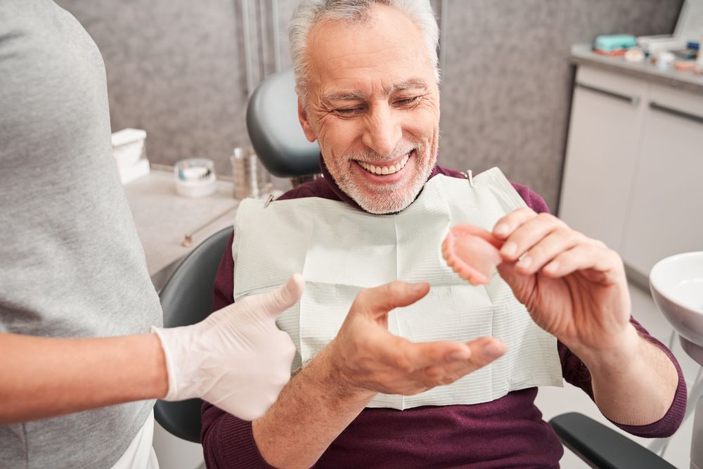 Older man smiling, holding dentures, dental office setting.