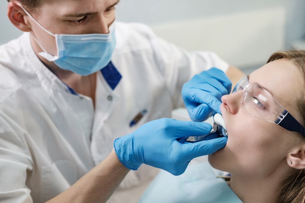 Dentist examining a patient's mouth, using tools and wearing mask/gloves, in a dental office.