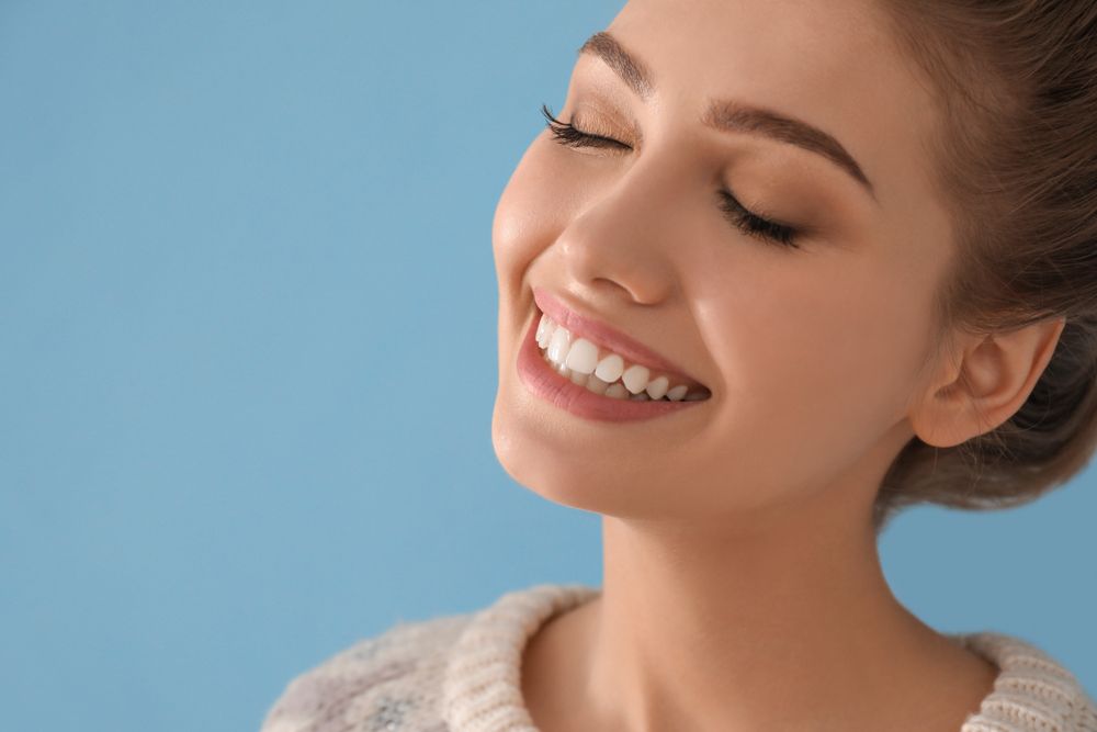 Woman with eyes closed, smiling widely, showing white teeth, against a blue backdrop.