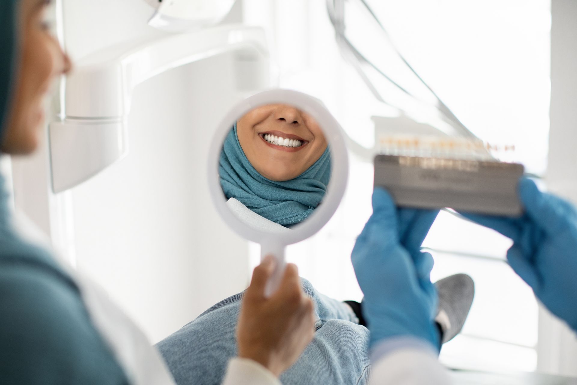 Woman smiles in dental chair, looking in mirror. Dentist holds shade guide, assessing teeth whiteness.