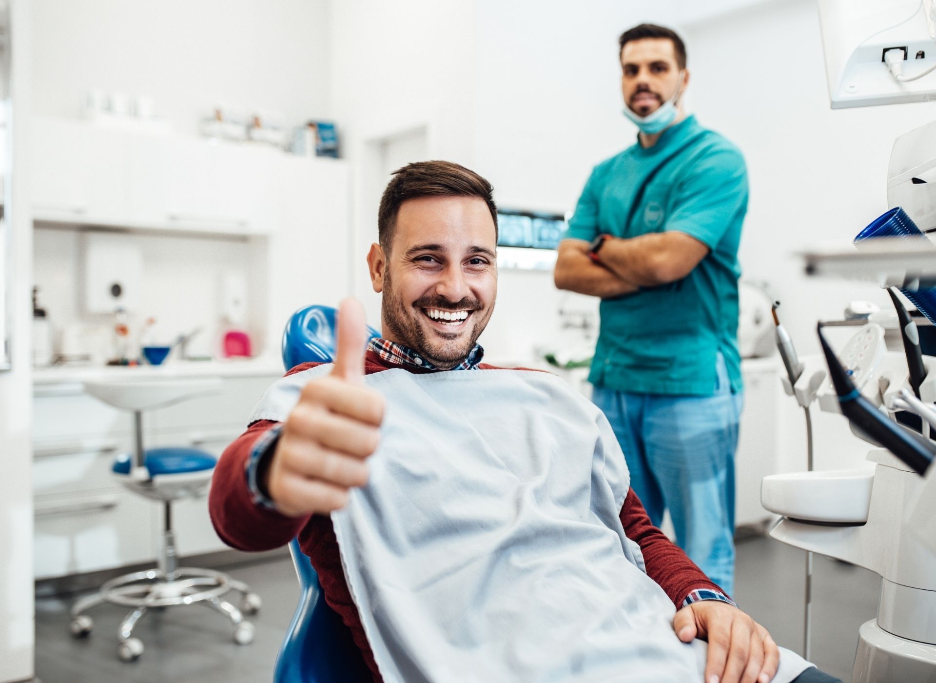Man in dentist chair gives thumbs up, smiling, with dentist in background. Bright, clean dental office.