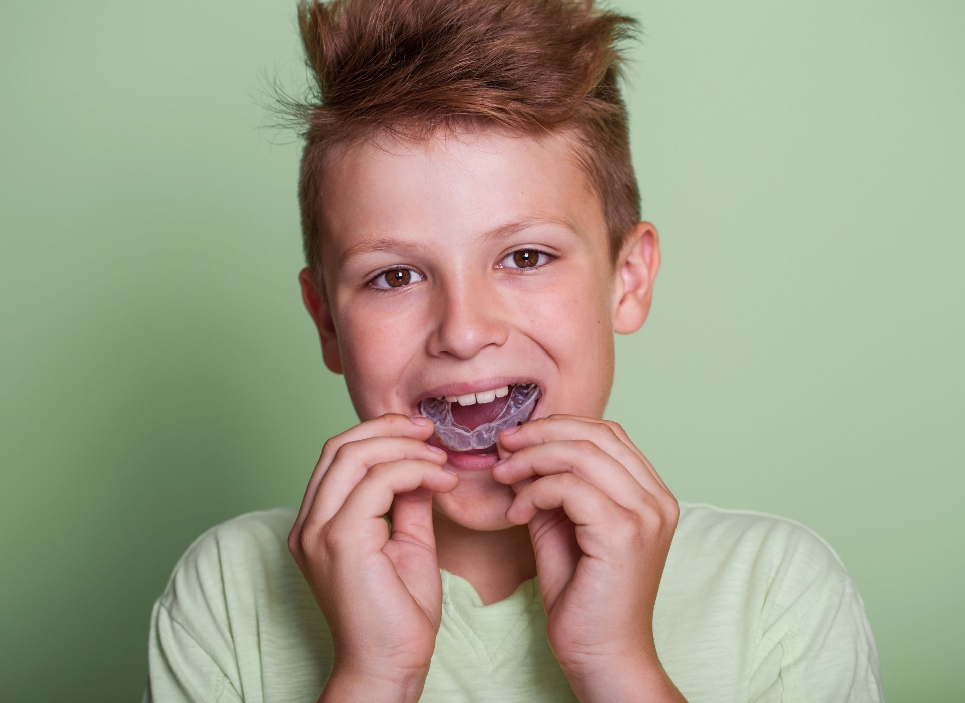 Boy placing a clear orthodontic retainer in his mouth against a green background.
