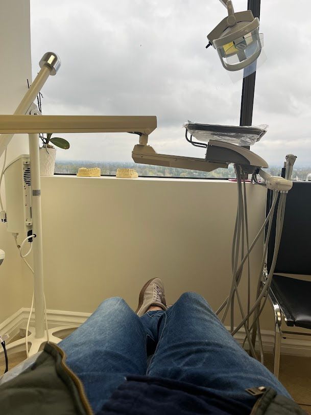 Farzad Dental- Person in a dentist chair with blue jeans, looking up towards dental equipment and a window with a cloudy sky.