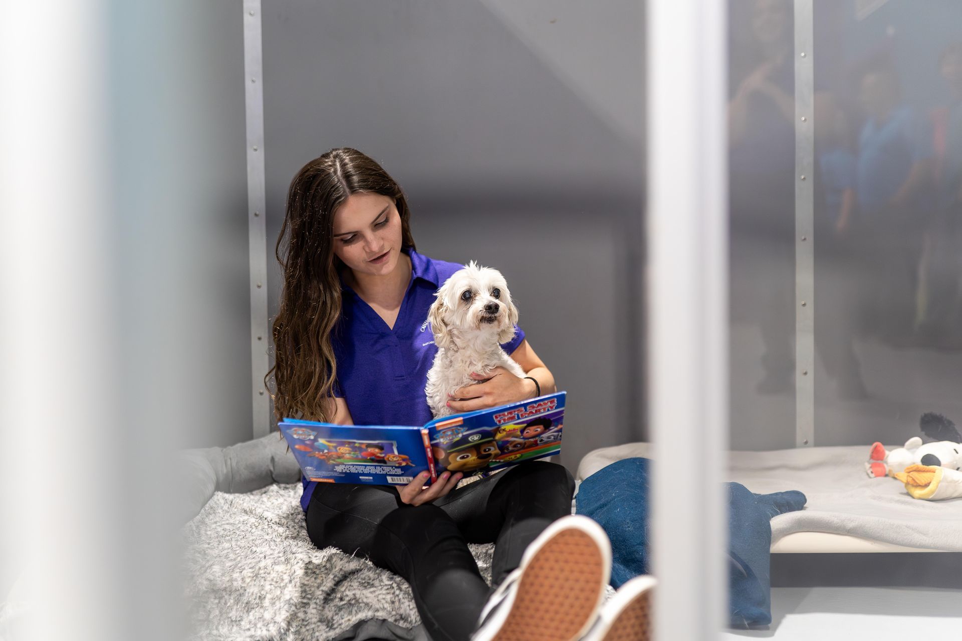 A woman is sitting on a bed reading a book to her dog.