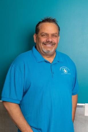 Man with dark hair, smiling, wearing a blue polo shirt, stands in front of a blue background.