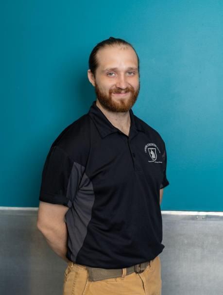 Man with beard in black shirt and tan pants, arms behind back, standing against a blue wall.