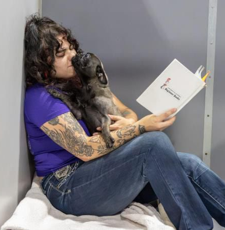 Woman with arm tattoos reads a book while hugging a puppy in a kennel; gray walls and a white towel are visible.