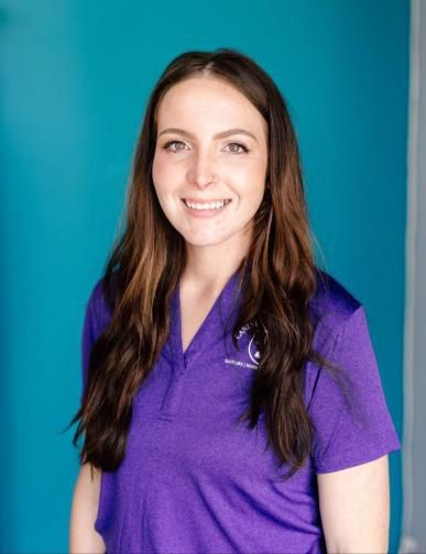 Woman with long brown hair smiles, wearing a purple shirt, in front of a teal wall.
