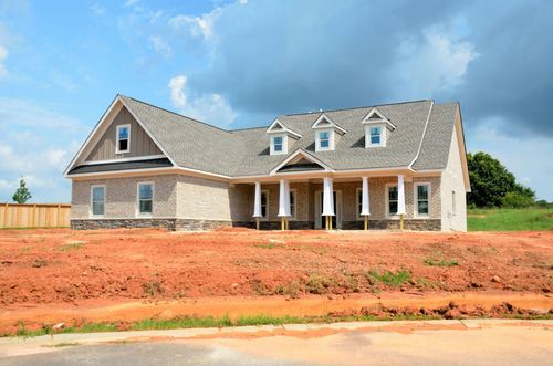 New house under construction with stone and siding exterior, front porch, and dirt lot under a blue sky