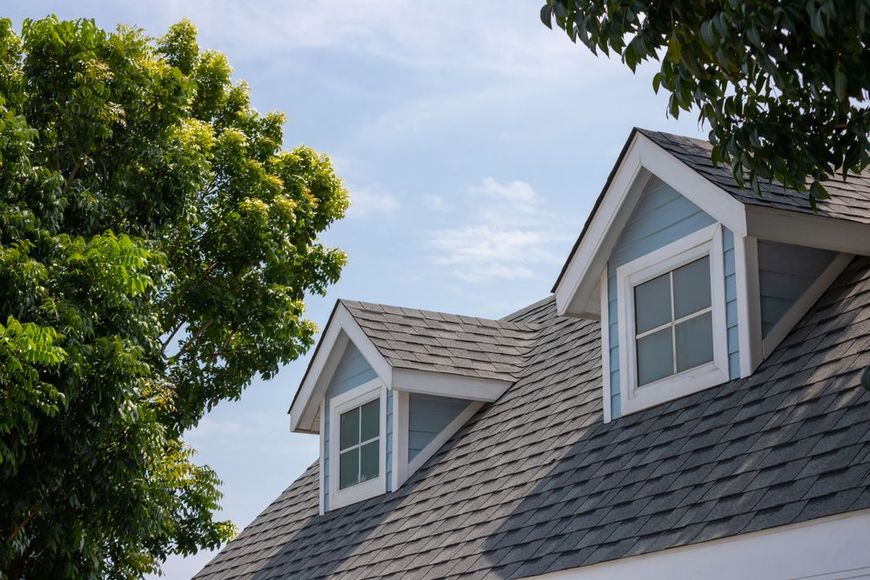 Gray-shingled house roof with dormer windows beside a leafy green tree under a blue sky