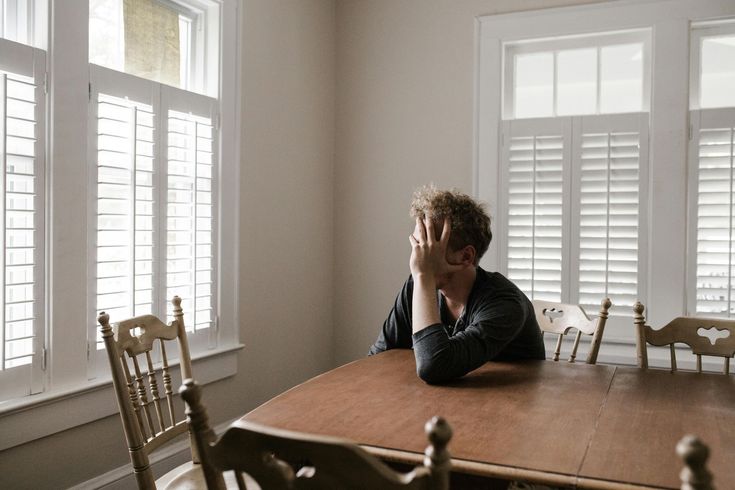 Person sitting at a wooden table in a bright room with large windows and white shutters