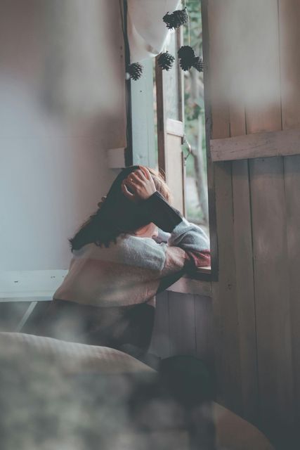 Person sitting by a window, resting their head on a pillow in soft indoor light.