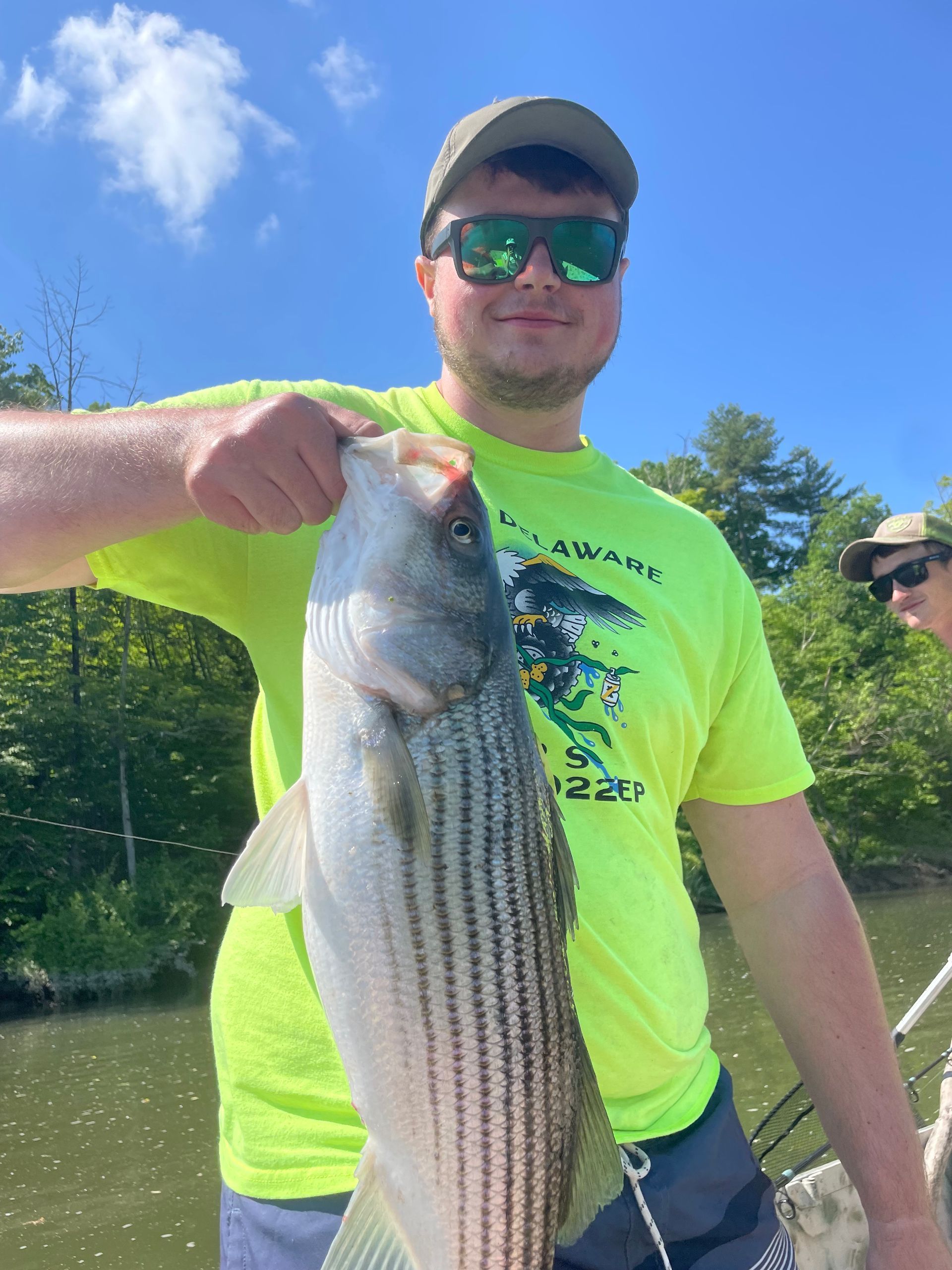 Ready Scout Aquatic Engineer, Andrew Hess,  in a bright yellow shirt and sunglasses holds a large striped bass while on a boat outdoors.