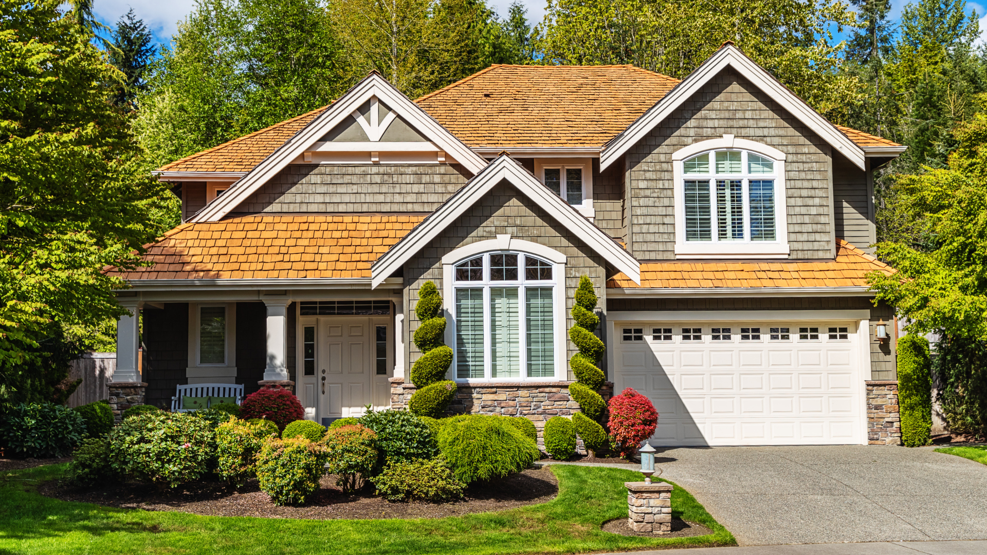 Two-story suburban house with a brown roof and gray siding, surrounded by green trees and bushes.