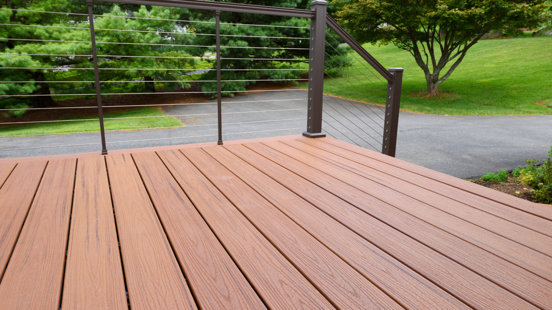 Brown composite deck with dark metal railing, overlooking a paved path and green lawn.