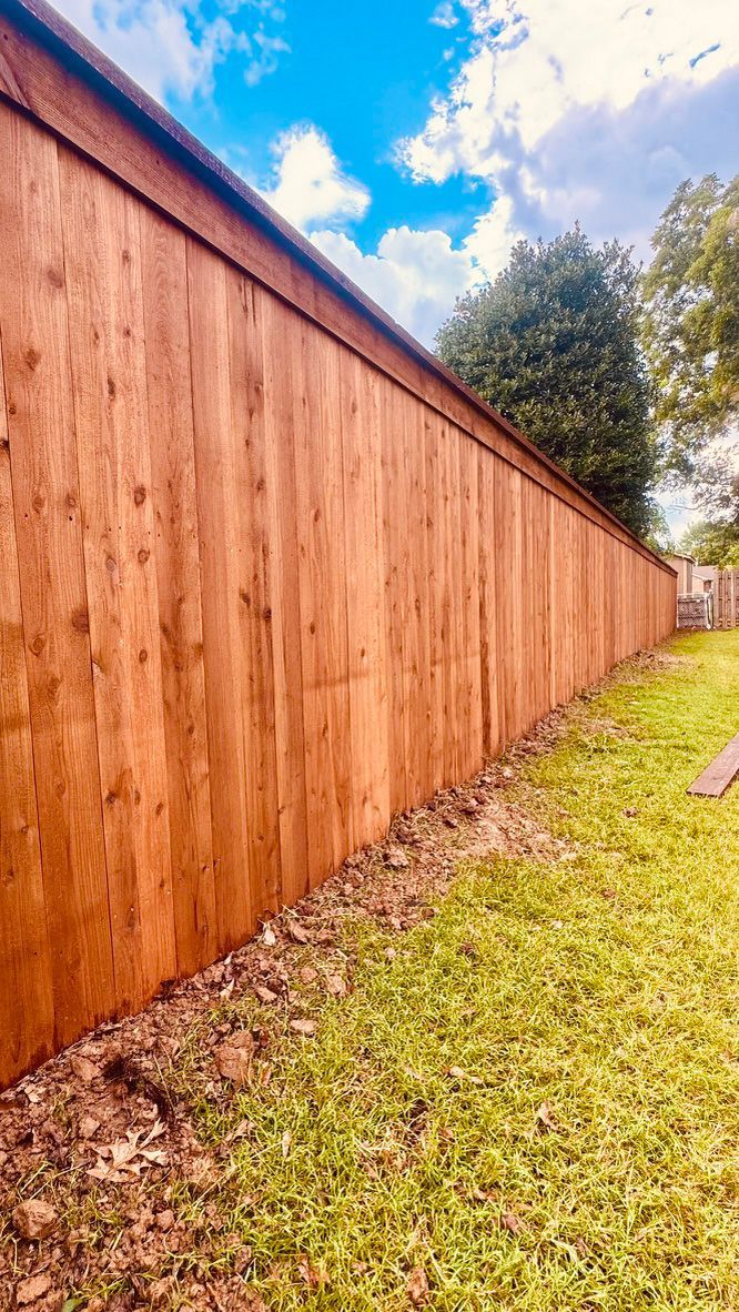 Wooden fence, stained brown, extends along a grassy yard under a blue sky with some trees.