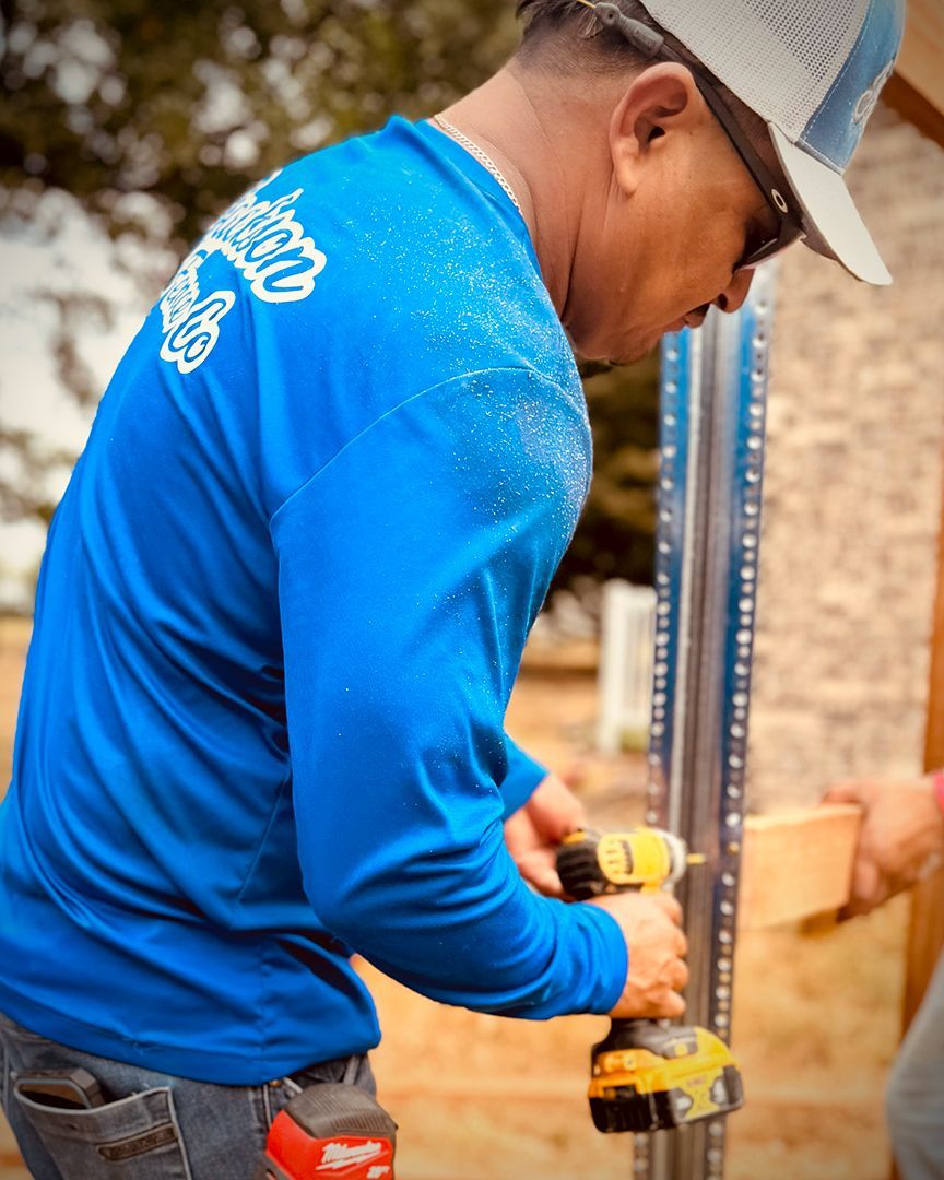 Person in blue shirt using a drill, working on metal structure outdoors. Sawdust visible.