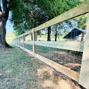 Wooden fence with wire mesh, in a grassy field, under trees. A small building is visible in the background.