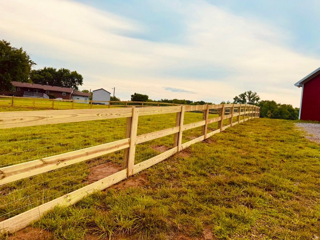 Wooden split-rail fence in a grassy field on a sunny day with a red barn visible.
