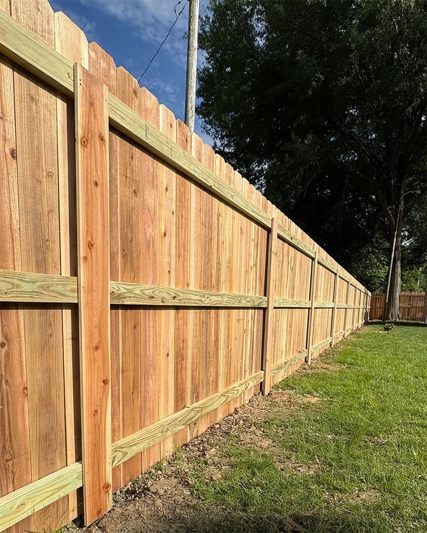 Wooden privacy fence with a scalloped top in a grassy yard, under a blue sky.