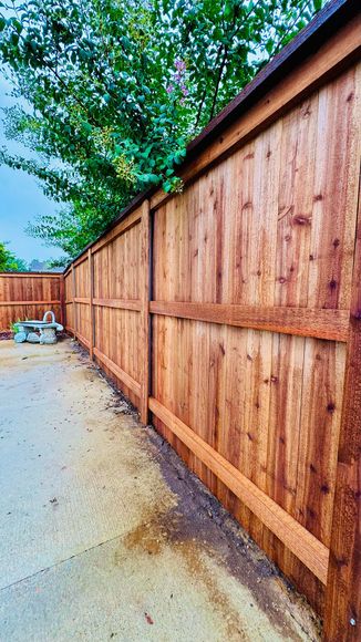 Wooden fence alongside a concrete path, under a cloudy sky with green foliage in the background.
