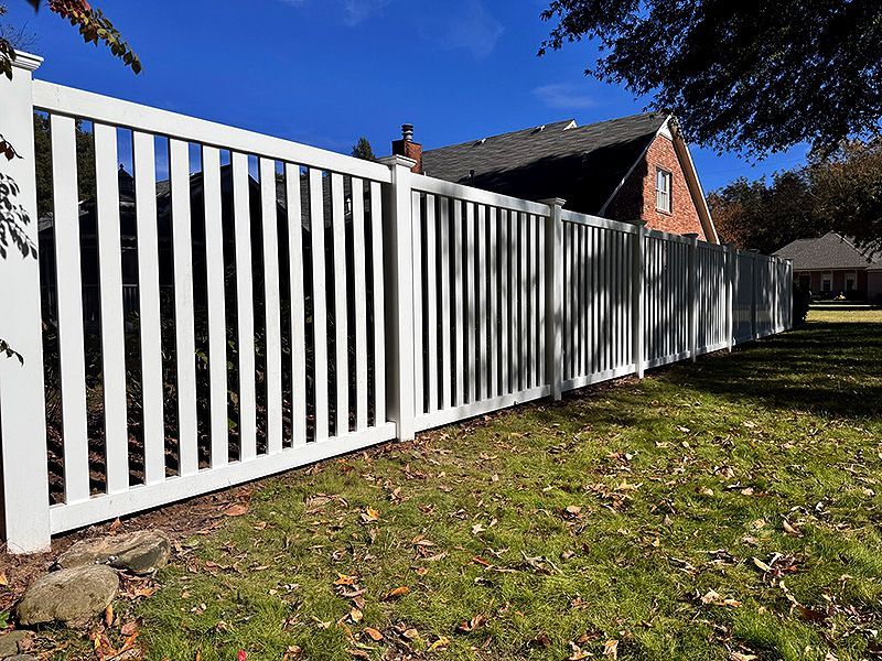 White picket fence along a grassy yard with a brick house and blue sky in the background