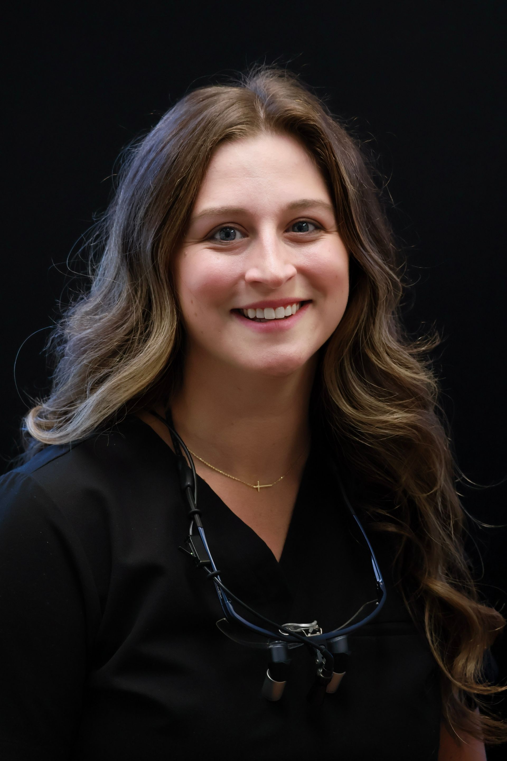 Woman in black scrubs smiles; loops and magnifying glasses hang around her neck.