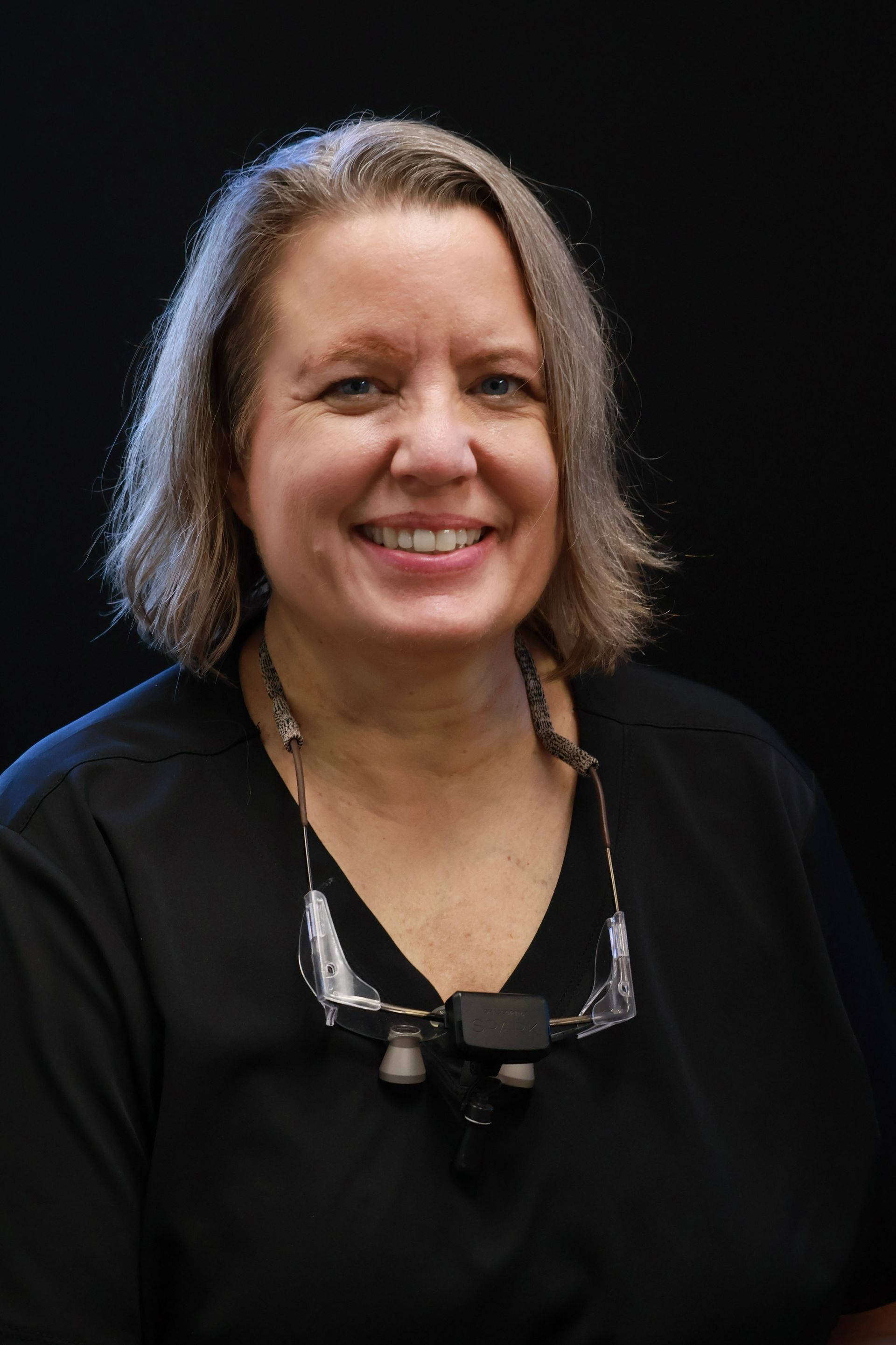 Woman smiling, wearing black scrubs and glasses around her neck, black background.