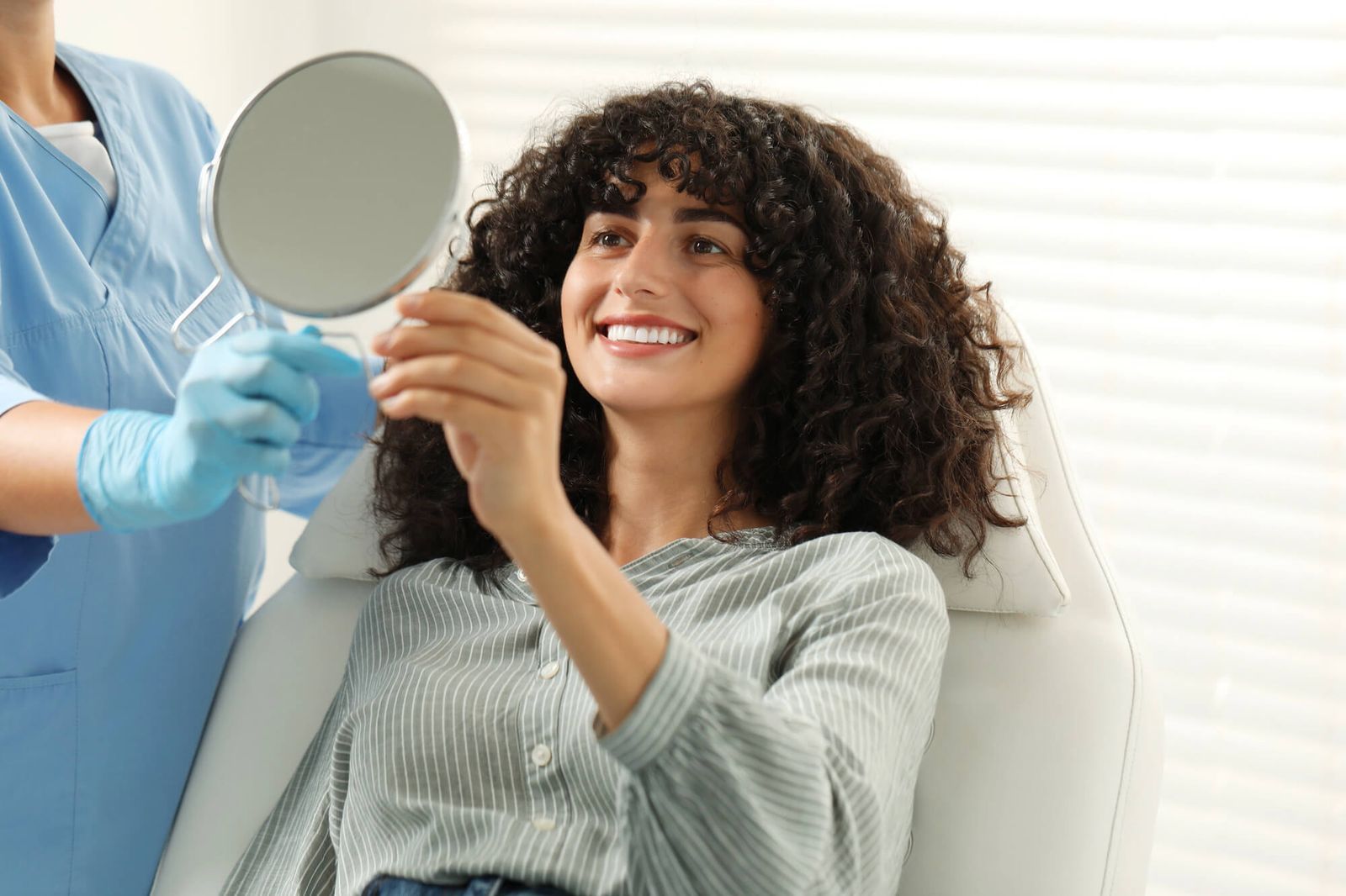 Woman smiling, looking at teeth in mirror held by gloved hand in a dental setting.