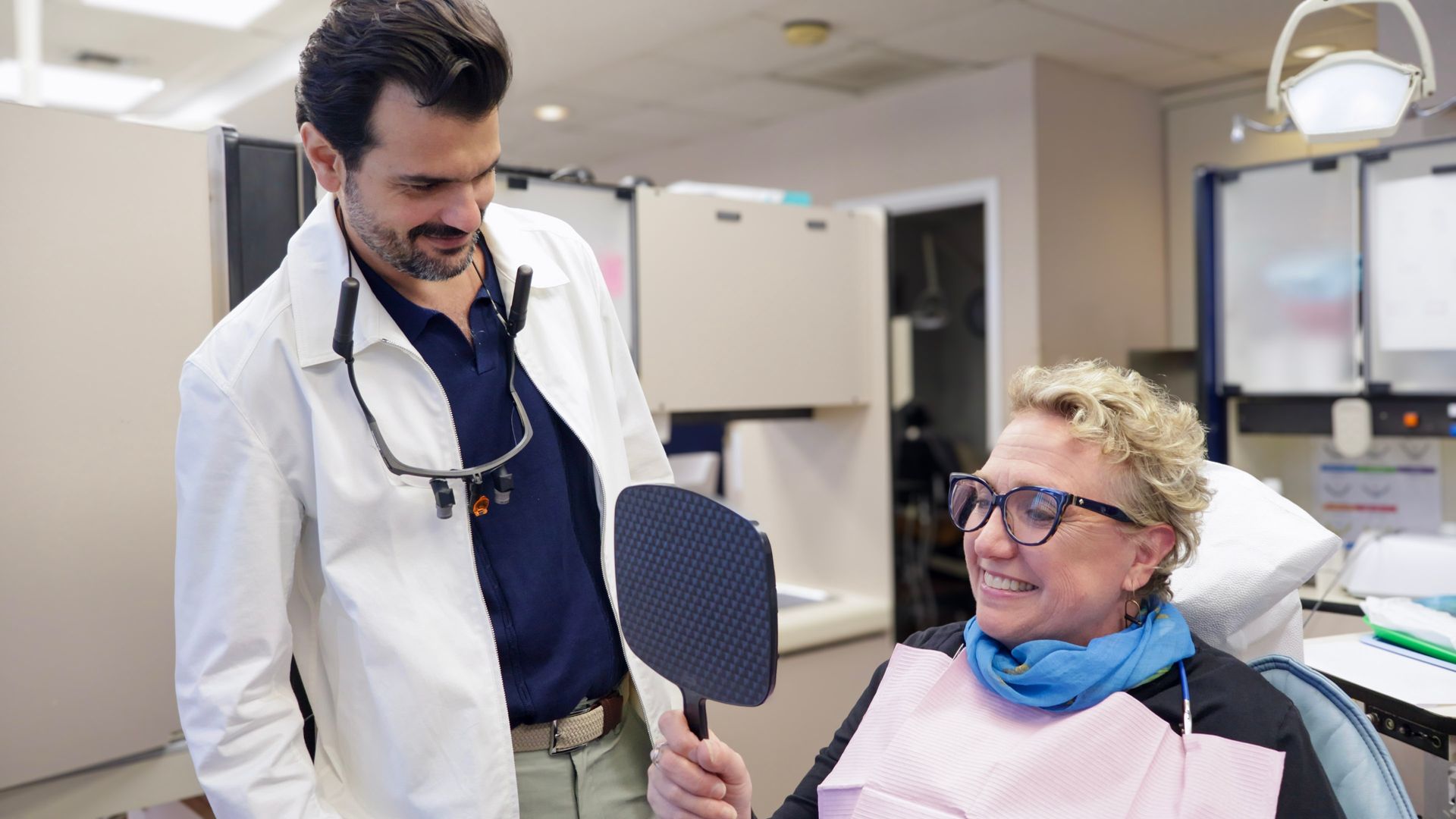 Dentist showing a patient their teeth in a dental office. The patient smiles, holding a mirror.