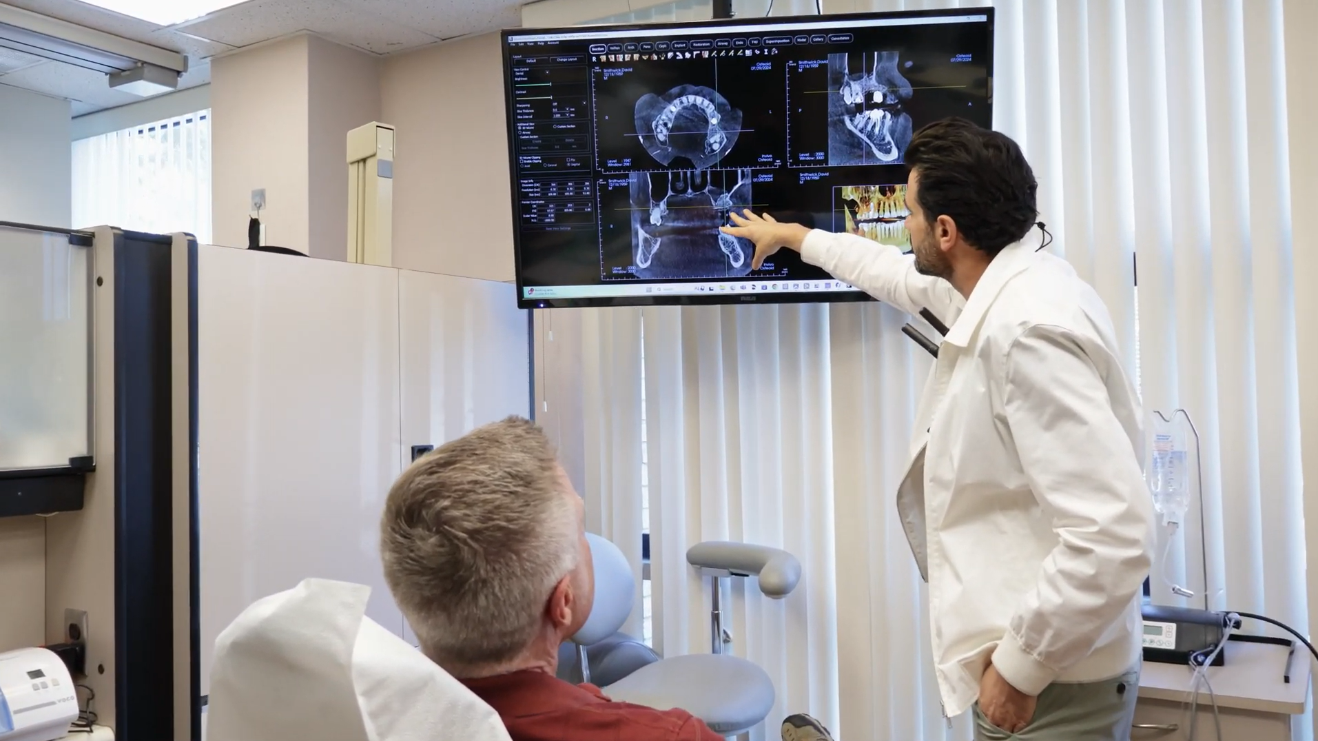 Dentist pointing at a dental scan on a large screen, explaining to a patient in a dental chair.
