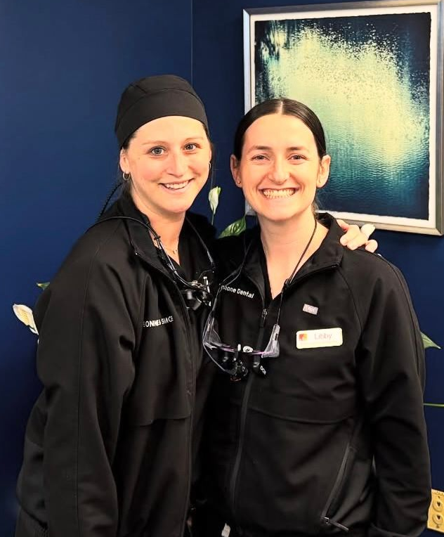 Two smiling dental professionals in black jackets and headwear, standing in front of a blue wall.
