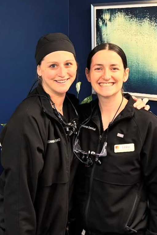 Two smiling dental professionals in black jackets and headwear, standing in front of a blue wall.
