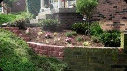 Brick retaining wall with tiered flower beds in front of a brick building and stairs. Lush green grass on the left.