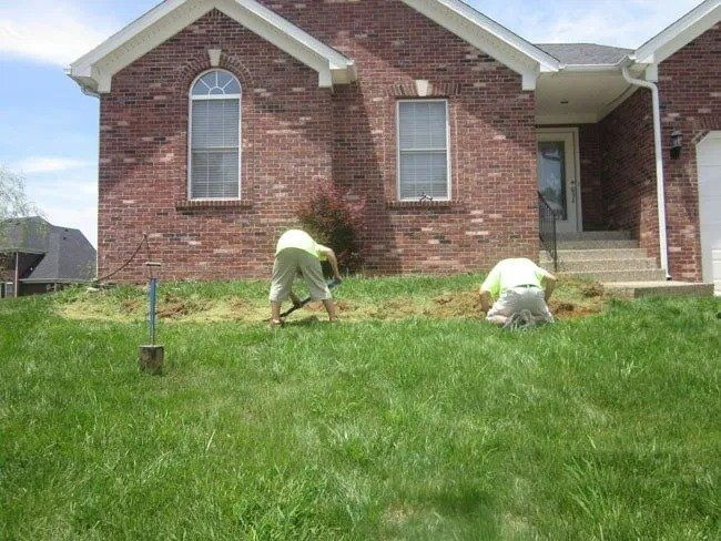 Two people in light-colored shirts and pants digging in a dirt patch in front of a brick house with a green lawn.