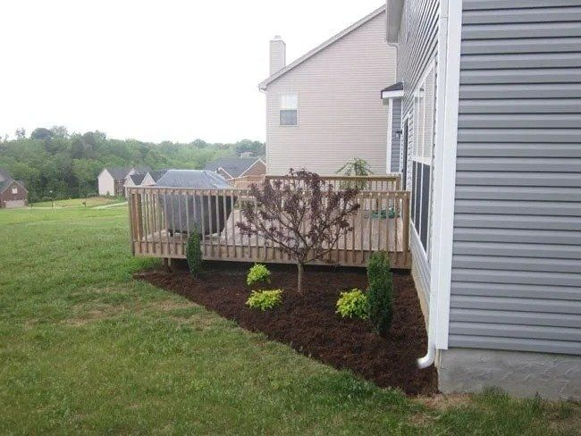 Backyard with a wooden deck and landscaped bed with small trees and shrubs next to a house with gray siding.
